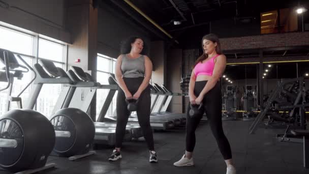 Dois adoráveis atletas do sexo feminino plus size agachados com kettlebells no ginásio — Vídeo de Stock