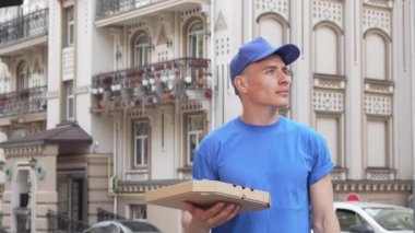 Neşeli pizza kuryesi şehir caddesinde etrafa baktıktan sonra kameraya gülümseyen