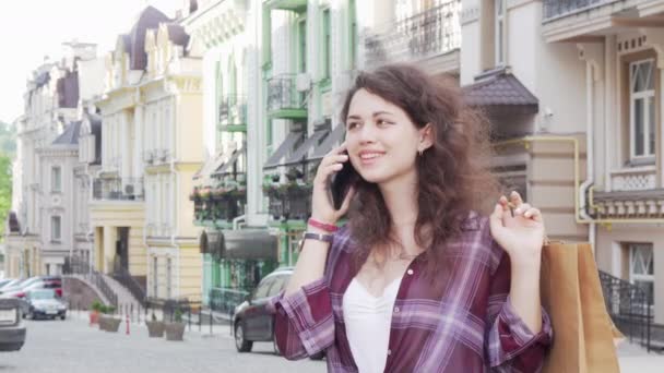 Urocza młoda kobieta rozmawia przez telefon na ulicy miasta trzyma torby na zakupy — Wideo stockowe