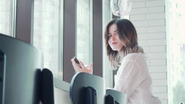 Mujer encantadora sonriendo a la amera usando el teléfono inteligente en la cafetería — Vídeo de stock