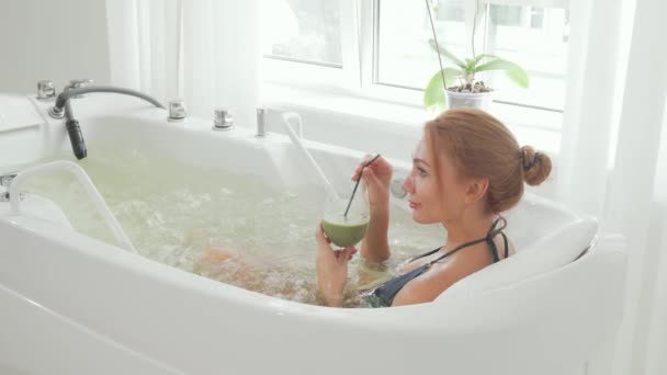 Vista trasera de una mujer bebiendo batido detox sentado en baño de hidromasaje — Vídeo de stock