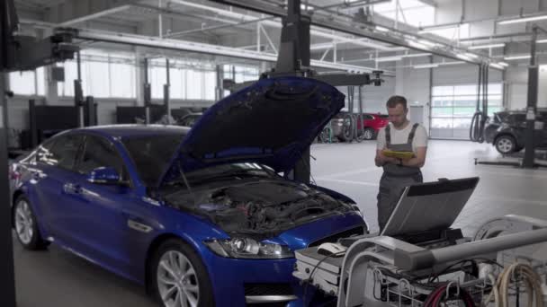 Зріла чоловіча механіка робить нотатки і використовує комп'ютер під час ремонту автомобіля — стокове відео