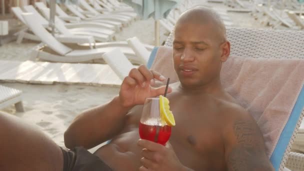 Bonito homem africano bebendo coquetel enquanto relaxa na praia — Vídeo de Stock