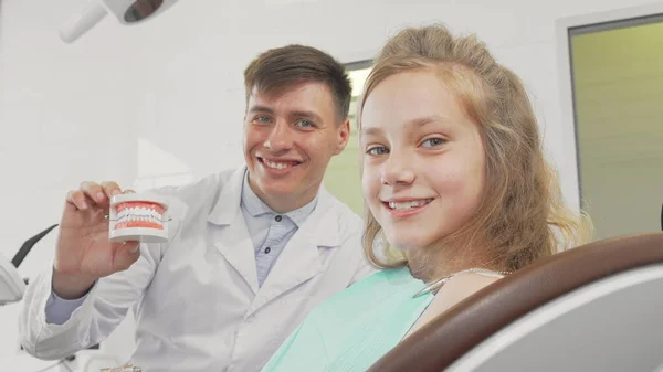 Ragazzina sorridente alla macchina fotografica dentista che tiene i denti modello sul retro — Foto Stock