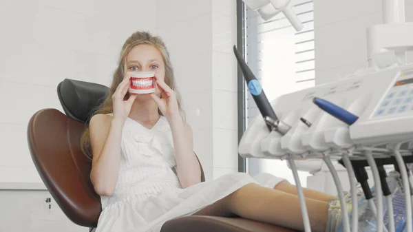 Красивая маленькая девочка улыбается держа модель челюсти сидя в стоматологическом кресле в клинике — стоковое фото