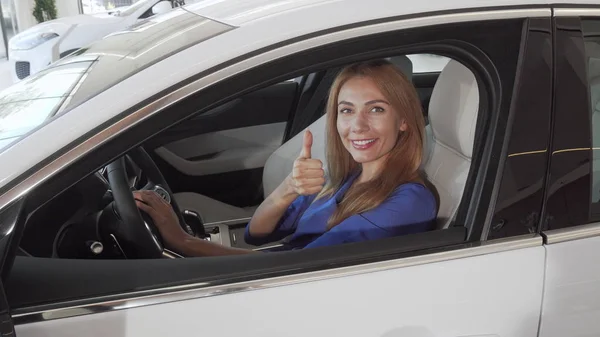 Hermosa conductora sonriendo a la cámara sentada en un coche nuevo en el concesionario — Foto de Stock