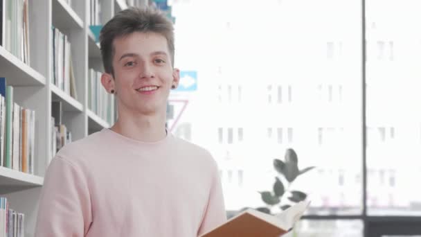 Veselý mladý muž se usmívá při čtení knihy v univerzitní knihovně — Stock video