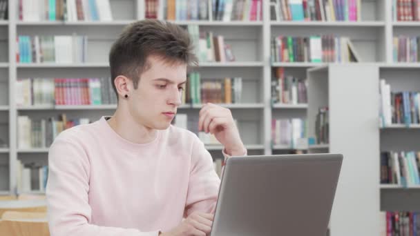Młody człowiek wygląda troskliwie podczas pracy nad laptopem w bibliotece — Wideo stockowe