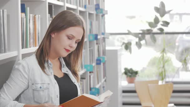 Schöne junge Frau lächelt in die Kamera, während sie in der Bibliothek liest — Stockvideo