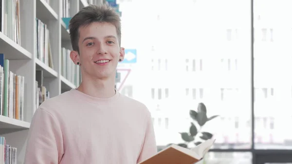 Веселый молодой человек улыбается, читая книгу в библиотеке колледжа — стоковое фото