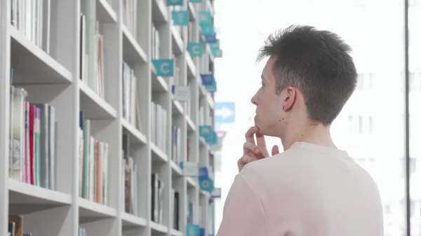 Young man looking thoughtfully at the bookshelves at the library — Stock Photo, Image