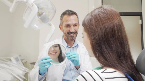 Vrolijke volwassen tandarts houdt een spiegel voor zijn vrouwelijke patiënt — Stockfoto