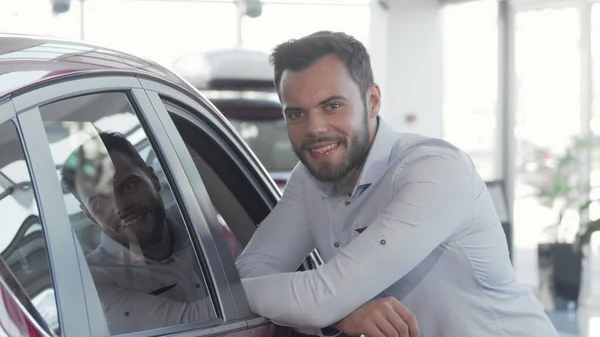 Handsome man smiling to the camera while examining car for sale at dealership