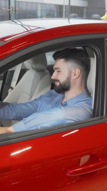 Zarif sakallı şık bir genç adam, galeride çarpan kırmızı bir arabanın sürücü koltuğunda otururken kendine güvenerek mutluluk saçar. Bu canlı sahne, yeni bir araç seçmenin coşkulu anını özetliyor.