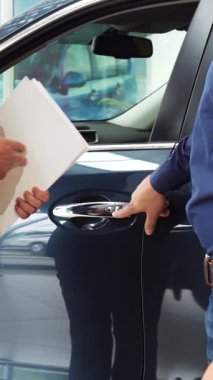 Modern bir araba galerisinde, yeni sahibi yeni arabasının sofistike kilitlerini hevesle inceliyor. İki iyi giyimli adam, biri açık mavi gömlekli, diğeri şık ceketli, sıkı bir tokalaşmayı sembolize ediyor.