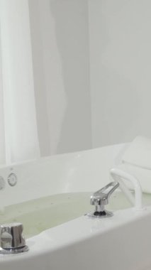 Işıltılı bir gülümsemesi olan neşeli bir kadın lüks bir girdap banyosuna adım atıyor ve rahatlatıcı bir hidromasaj yaptırmaya hazır. Pencereden süzülen yumuşak doğal ışıkla aydınlatılan sakin spa ortamı davetkar bir davet yaratıyor.