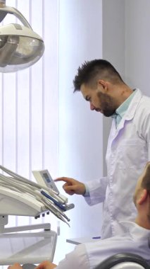 Kendini adamış beyaz önlüklü esmer bir erkek dişçi şık, modern bir diş sandalyesinde rahat bir şekilde oturan sarışın beyaz hastasıyla dikkatlice etkileşime girer. Parlak, havadar ofis, yumuşak doğal ışıkla aydınlatılır.