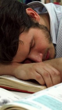 Yorgun, esmer, beyaz bir erkek öğrenci, dağınık bir kütüphane masasında uyukluyor, kafası ders kitaplarının arasında açık bir kitap üzerinde dinleniyor. Yakın plan, gece geç saatlere kadar çalışmanın yorgunluğunu yansıtan huzurlu ifadesini yakalıyor.