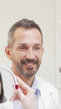 Neşeli, olgun bir dişçi kadın hastası parlak gülümsemesini parlak bir aynada izlerken gururla ışıldıyor. Bu iç açıcı an, diş bakımının neşesini özetliyor. Parlak ve modern bir klinikte güven ve memnuniyeti vurguluyor.