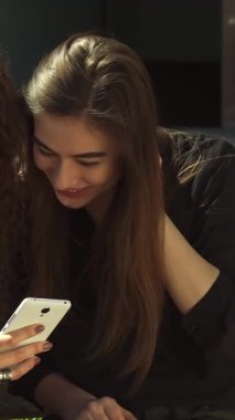 Şık siyah tişörtlü neşeli bir beyaz kız, her ikisi de güneşli bir açık hava bankında oturan kıvırcık saçlı arkadaşıyla cep telefonunda heyecan verici bir keşfi paylaşıyor. Işıl ışıl gülüşleri ve etraflarındaki gür yeşillik