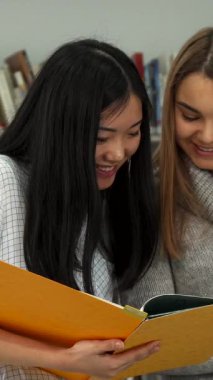 Renkli kitaplarla dolu canlı bir kütüphanede, biri parlak siyah saçlı iki hevesli kız öğrenci ve diğerinin uzun esmer saçı var, hevesle notlarından yola çıkarak görüşlerini paylaşıyorlar. Parlak gülümsemeleri ve hareketli ifadeleri derin bir şeyi yansıtıyor.