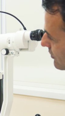 Odaklanmış bir adam dikkatle yüksek teknolojili bir göz inceleme cihazına bakar. Parlak, steril bir ofiste modern tanıların hassasiyetini gösterir. Atmosfer profesyonellikle dolu. Göz doktoru vizyonunu sağlama almaya hazırlanıyor.