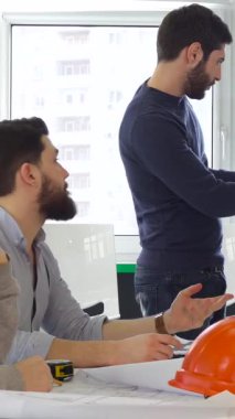 Canlı genç bir mimar takımı, parlak ve modern bir ofiste yenilikçi bir yapı projesi hakkında ateşli bir tartışmaya girişiyor. Kendinden emin, esmer, sakallı bir adam parmak izi çizelgesinin yanında duruyor. Karmaşık planlara işaret ediyor.