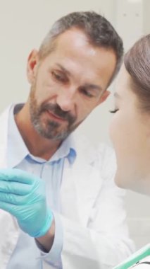 Neşeli bir kadın, dişçi koltuğunda otururken kendine güveni yayar. Parlak gülüşü odayı aydınlatır. Kıtır önlük giyen ilgili dişçi detaylı bir diş modelini elinde tutarak, güveni vurgulayan sıcak ve profesyonel bir atmosfer yaratıyor.