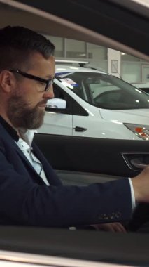 Özel dikim takım elbiseli dengeli bir satış müdürü modern bir araçta rahatça oturan olgun, sakallı bir adamla özellikleri tartışmak için öne eğilir. Sahne, arabaların ileri teknolojilerini keşfederken heyecan ve profesyonellik yayıyor.