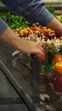 Yakın çekim, canlı bir süpermarkette parlak kırmızı ve güneşli sarı dolmalık biberler arasındaki seçenekleri zarifçe tartan bir adamın elini yakalar. Sahne taze ürünlerle dolup taşıyor, canlı sebze renklerini ve dokularını gözler önüne seriyor,