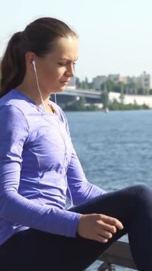 Kararlı genç beyaz bir kadın, ışıldayan bir nehir yürüyüşü boyunca bacaklarını açıyor, enerji egzersizi için hazırlanıyor. Uzun siyah saçları parlak bir at kuyruğuyla, kulaklıklarla en sevdiği müziği dinliyor, düşünceli bir şekilde bakıyor.