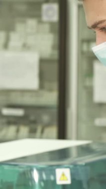 Görüntü, test sonuçlarına dalmış beyaz önlüklü odaklanmış genç bir laboratuvar teknisyenini gösteriyor. Lateks eldivenler ve koruyucu bir maske takarak bulgularını özenle sarı bir klasöre kaydediyor.