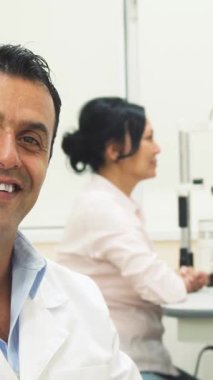 Kendine güvenen, şık, mavi çerçeveli gözlüklü bir erkek doktor, olumlu ve kendinden emin bir ışık saçıyor. Arka planda, kendini işine adamış bir kadın doktor bir kadının gözlerini dikkatlice inceleyerek, sıcak ve profesyonel bir atmosfer yaratıyor.