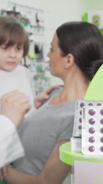 Parlak ve neşeli bir eczanede, kendini işine adamış bir eczacı renkli bir ilaç şişliğini küçük bir çocuğa uzatırken şefkatli annesi gururla izliyor. Bu iç açıcı sahne, aile sağlığının ve desteğin önemini vurguluyor.