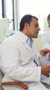 Güneşli bir muayenehanede, beyaz önlüklü iki erkek doktor X-Ray görüntülerini titizlikle incelerken, genç bir kadın umut dolu bir ifadeyle sonuçlarını bekliyor. Bu sahne adanmışlığı güzel bir şekilde gösteriyor.