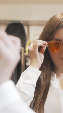 Çekici genç bir kadın ışıl ışıl bir butikte şık güneş gözlüğü denerken neşe saçıyor. Aynada yansıması parlıyor. Göz giysisinin neşeli renkleri şık kıyafetini tamamlıyor. Alışverişi zevkli bir maceraya dönüştürüyor.