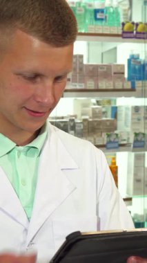 Gıcır gıcır beyaz bir tıbbi elbise giymiş odaklanmış bir erkek eczacı, tablet üzerinde gezinirken bir telefon konuşmasını ustalıkla yapıyor ve hasta bakımına olan bağlılığını gösteriyor. Düzenli ve canlı bir dizi ilaç ürünüyle çevrelenmiş bir şekilde,