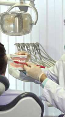 Parlak, modern bir diş kliniğinde, yetenekli bir esmer erkek dişçi, insan çenesinin detaylı bir modelini kullanarak doğru diş fırçalama tekniğini tutkuyla gösterir. İlgili genç erkek hastası dişçinin ustalıkla manevra yapmasını yakından izliyor.
