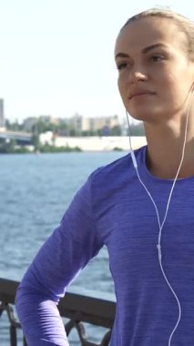 Genç beyaz bir kadın kollarını uzatıyor, neşe dolu bir gülümsemeyle etrafa bakıyor nehir kenarındaki canlı manzarayı kucaklıyor. Parlak menekşe bir bluz giyip mp3 çalarıyla müzik dinliyor. Ona hazırlanırken enerji ve kendine güven saçıyor.