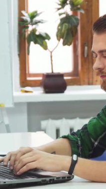 Siyah saçlı ve sakallı neşeli genç bir adam parlak beyaz bir masada oturuyor ve güneşli bir sınıfta dizüstü bilgisayarında yazı yazıyor. Sıcak gülüşü ve rahat tavırları, canlı yeşil bitkiler ve doğal ışıkla güçlendirilmiş davetkar bir atmosfer yaratıyor.