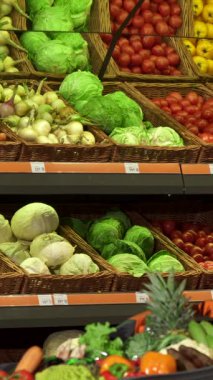 Odaklanmış genç beyaz bir adam canlı bir hipermarkette parlak kırmızı bir dolma biber ve gevrek yeşil bir lahana da dahil olmak üzere canlı sebzeler seçiyor. Taze ürünlerden oluşan bir kaleydoskop ile çevrelenmiş, sağlıklı yaşamın heyecanını somutlaştırıyor.