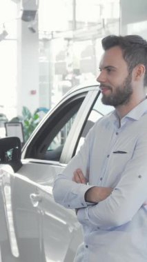 Kendine güvenen genç beyaz bir adam parlak, parlak yeni aracının yanında duruyor, elinde parlak bir gülümsemeyle arabanın anahtarlarını tutuyor. Parlak, modern sergi salonu, verimli yeşillik ve çarpıcı kırmızı bir fiyonk ile, heyecan ve yeni sahiplik sevinci yayar.