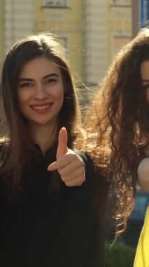 Dalgalı saçları ve ışıl ışıl gülümsemeleri olan iki zarif genç kadın, canlı bir şehir zeminine karşı coşkulu başparmaklarını uzatarak alışveriş çantalarını gururla gösteriyorlar. Neşeli ifadeleri ve şık kıyafetleri özlerini yansıtıyor.