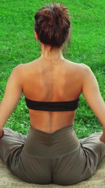 Genç bir kadın doğal yoga minderinin üzerinde huzur içinde oturur, sırtını izleyiciye çevirir ve nazikçe yoga pozu verir, ellerini meditasyon hareketine sıkıştırır. Etrafı canlı yeşil otlarla çevrili, rahat duruşu ve sakin ifadesi barışı yayıyor.