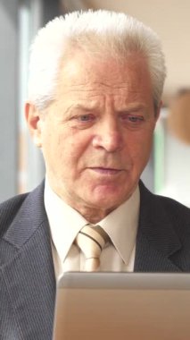 Özel dikim takım elbise ve kravat giymiş zarif bir kıdemli işadamı, şık bir kafede dizüstü bilgisayarı üzerinde çalışırken dumanı tüten bir fincan kahveden zevk alıyor. Seçkin beyaz adam, çarpıcı gri saçlar ve odaklanmış bir ifadeyle,