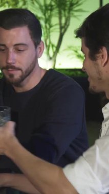 İki şık erkek arkadaş viski bardaklarını şen şakrak bir tostla tokuşturuyorlar, yemyeşil ve çevre aydınlatmalarıyla çevrili. Parlak bar atmosferi sıcaklığı ve dostluğu yansıtıyor, yoğun bir yoğunluktan sonra dostluğun ve gevşemenin özünü yakalıyor.