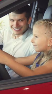 Işıl ışıl sarı saçları saçlarıyla, parlak küçük bir kız babasının yanında, parlak kırmızı bir arabayla direksiyon sallıyormuş gibi yapıyor. Bulaşıcı gülüşleri ve şakalaşmaları aile neşesi ve bağlantıları için doyurucu bir tablo oluşturuyor.