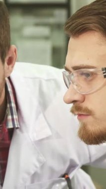 Yakın plan çekimde iki beyaz önlüklü genç erkek laboratuvar teknisyeni ciddi bir tartışmaya dalmış, yoğun ifadeleri analizlerinin ağırlığını yansıtıyor. Etrafı son teknoloji ekipmanlarla çevrili ve...