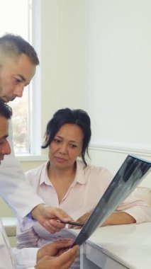 İyi ışıklandırılmış bir muayenehanede endişeli bir ifadeye sahip genç bir kadın dikkatle, biri ciddi ve odaklanmış iki doktor olarak röntgen sonuçlarını tartışıyor. Sahne profesyonellik ve empati gösteriyor, netliğin önemini vurguluyor.