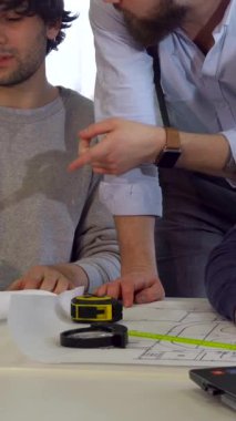 Parlak, modern bir çalışma alanında, üç Kafkasyalı mimar planlar ve proje belgeleriyle çevrili ruhsal bir tartışmaya giriştiler. Gıcır gıcır beyaz gömlekli, sakallı esmer bir adam öne eğiliyor ve hareketli bir şekilde koltuğuna rehberlik ediyor.